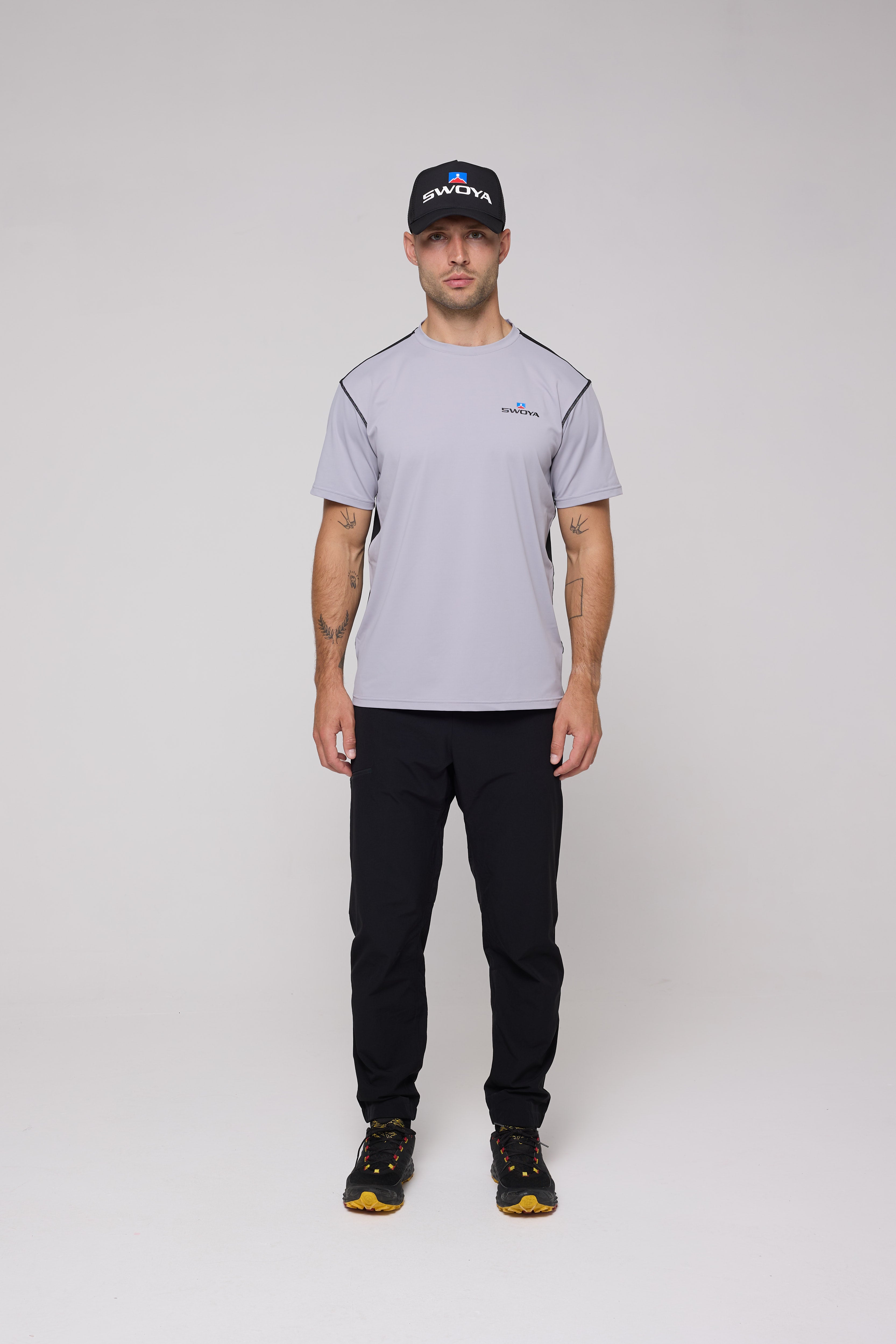 Man wearing a gray t-shirt, black pants, and a black cap with a logo on a white background